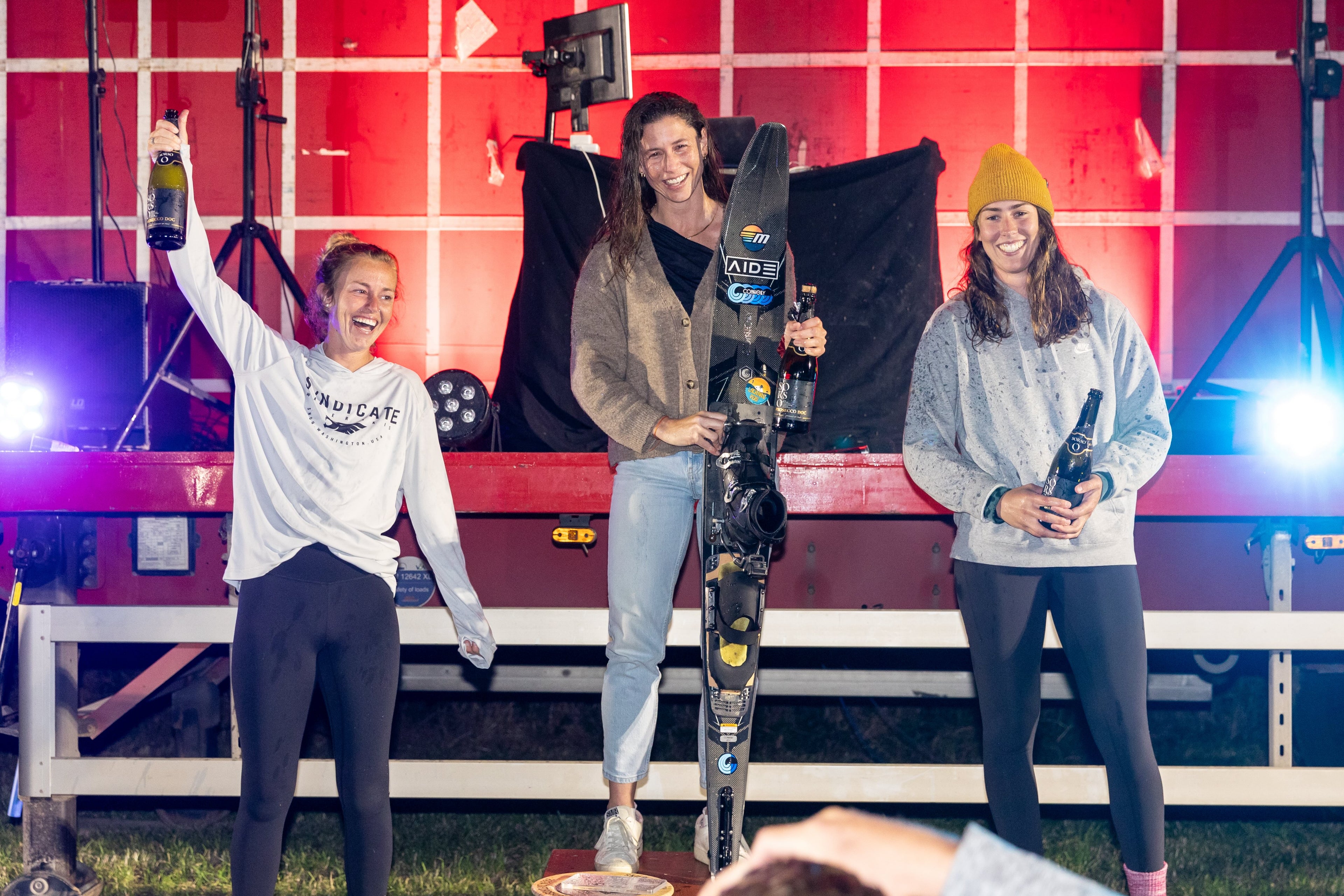 Three female athletes stand on a podium holding bottles and a water ski, smiling in front of a red-lit stage.