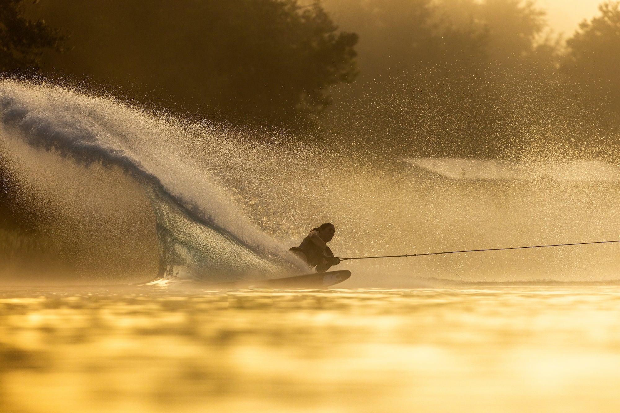 A skier carves through golden water at sunrise, creating a dramatic spray with their silhouette in the light.