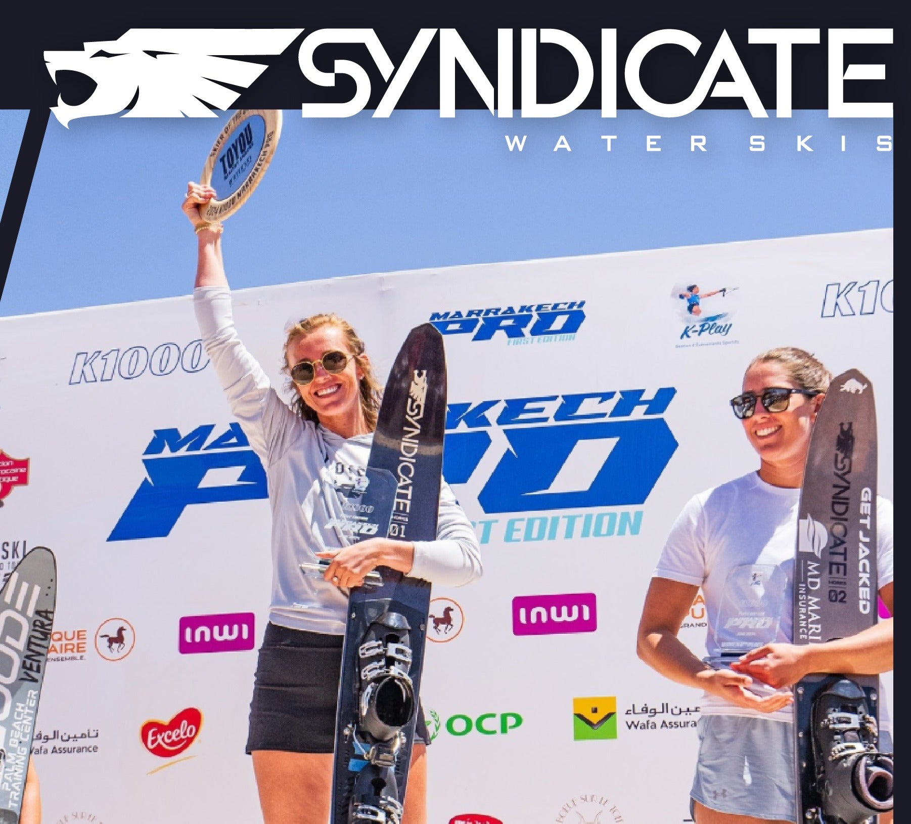 A female skier holds a plate trophy and a black Syndicate ski. Other competitors stand beside her with event banners behind.