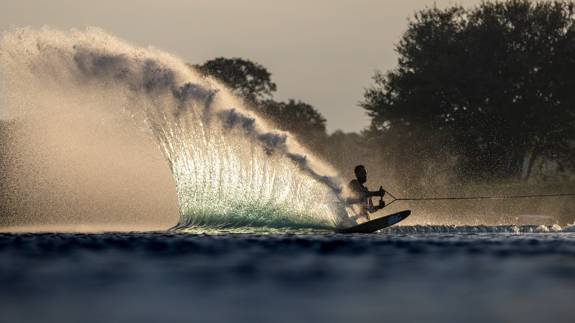 Water Skier throwing up a huge slash that is glowing in the sun riding a HO Sports ski and wearing a HO Sports Life Jacket.